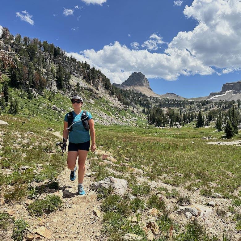A woman wearing a running vest walks along a trail with mountains in the background
