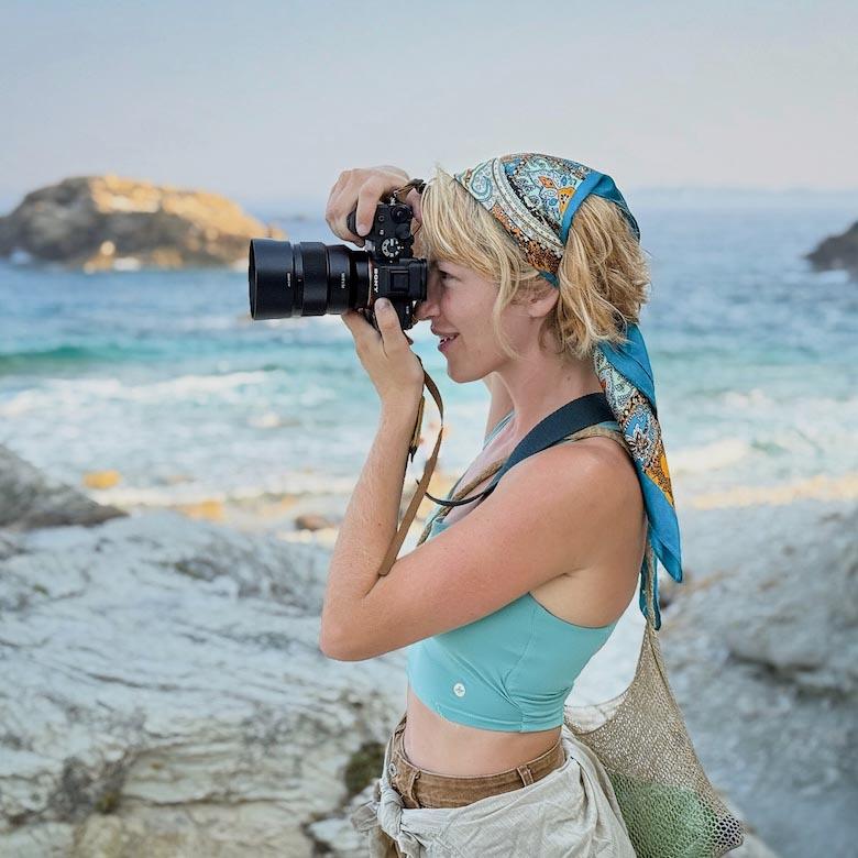 A woman standing next to the ocean looks through a camera lens