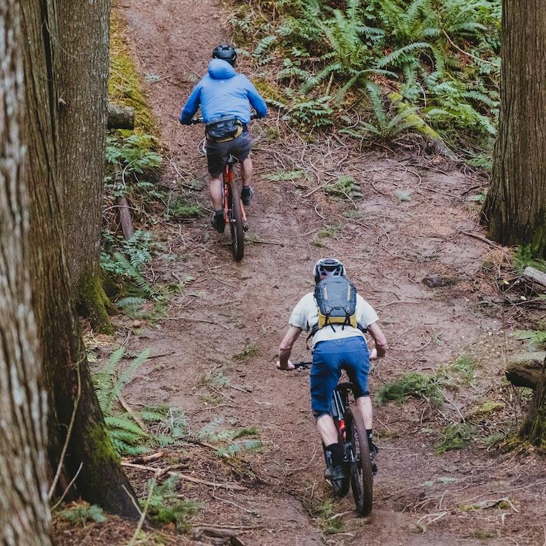 Two bikers ride up a mountain bike trail wearing backpacks