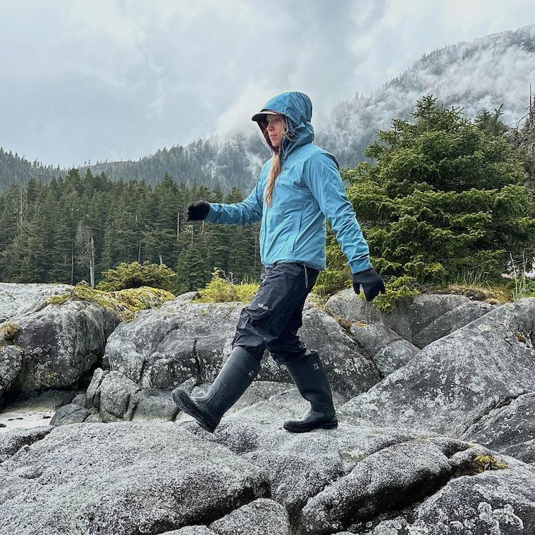 A woman in rain boots and rain jacket walks across rocks on a rainy day