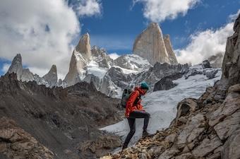 A woman hikes up steep rocks with jagged mountains and clouds in the background